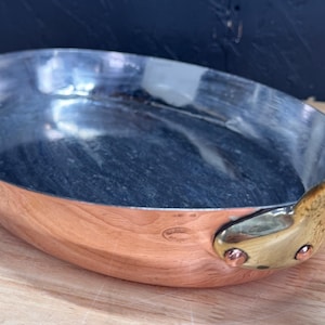 Op de afbeelding: Een koperen koekenpan met een messing handvat. De binnenkant is een donker, anti-aanbak oppervlak. De pan staat op een houten oppervlak, met een donkerblauwe achtergrond.