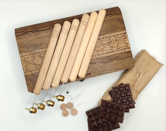 May include: A selection of wood-handled tools with brass tips, displayed on a decorative wooden box. The tools are alongside small wooden circles, needles, and dark chocolate bars. A wrapped package is also included.