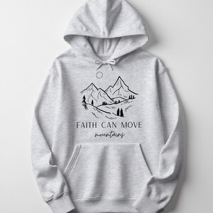 Könnte beinhalten: Ein hellgrauer Kapuzenpullover mit einer Vordertasche und Kapuze mit Kordelzug. Auf der Vorderseite befindet sich eine schwarze Strichzeichnung von Bergen, Bäumen und einer Sonne mit dem Text "Faith can move mountains" in Schreibschrift.