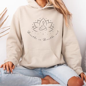 Könnte beinhalten: Ein beiger Hoodie mit einem schwarzen Lotusblüten-Design und dem Text "Breathe in Breathe out".