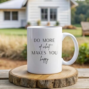 Könnte beinhalten: Weiße Keramiktasse mit dem Text "DO MORE of what MAKES YOU happy" in schwarzer Schrift. Die Tasse steht auf einem runden Holzuntersetzer mit natürlichem Rand. Im Hintergrund ein weißes Haus und Grün.