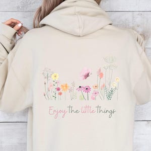 Könnte beinhalten: Beige Kapuzenpullover mit floralem Design, darunter ein rosa Schmetterling und der Schriftzug "Enjoy the little things". Das Design zeigt eine Vielzahl bunter Blumen und Blätter und schafft eine skurrile, von der Natur inspirierte Ästhetik.