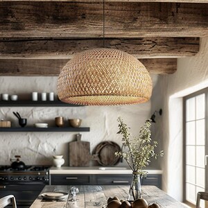 Op de afbeelding: Een rustieke houten eettafel met een geweven rieten hanglamp erboven. De tafel is gedekt met borden en glazen, en er is fruit in een kom op de tafel. De tafel is omringd door stoelen, en de kamer heeft een rustieke, boerderij-achtige sfeer.
