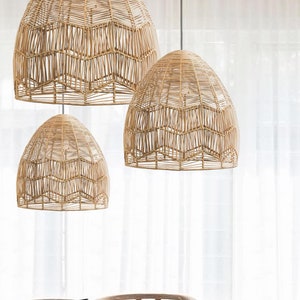 May include: A dining room with a large, round wooden table and woven chairs. Three woven pendant lights hang above the table. A potted tree is in the corner, and a sheer white curtain is in the background. The room has a natural, rustic feel.
