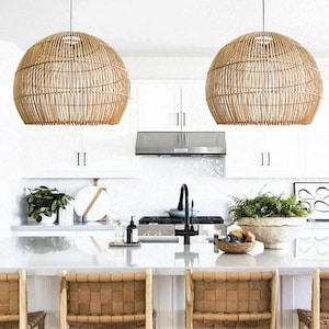 May include: Two large, dome-shaped pendant lights crafted from natural light brown woven rattan. Each fixture features a vertical slatted design with a circular top mount, suspended by thin black cords above a kitchen island with woven chairs.