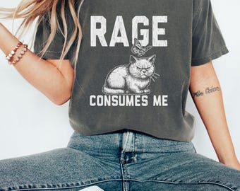 Puede incluir: Camiseta gris oscuro con el texto blanco "RAGE CONSUMES ME". Debajo del texto hay un gato de dibujos animados con expresión enfadada, con una mariposa encima de su cabeza. La camiseta es de estilo casual.