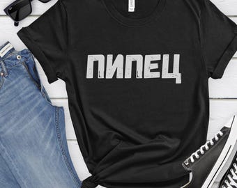 May include: Black t-shirt with the Russian word "ПИПЕЦ" printed in white on the front.