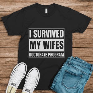 May include: Black t-shirt with the text "I SURVIVED MY WIFES DOCTORATE PROGRAM" in white. The shirt is paired with blue jeans and white canvas sneakers on a wooden surface.