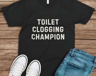 May include: Black t-shirt with the text "TOILET CLOGGING CHAMPION" in white. The shirt is paired with light blue jeans and white canvas sneakers. The items are arranged on a wooden surface.