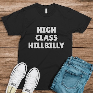 May include: Black t-shirt with the words "HIGH CLASS HILLBILLY" in white block letters. The shirt is paired with blue jeans and white Converse sneakers, all arranged on a wooden surface.