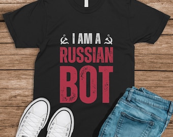 May include: Black t-shirt with the text "I am a Russian Bot" in red and white. The text is stylized with a distressed look. The shirt is worn over a pair of blue jeans and white sneakers.