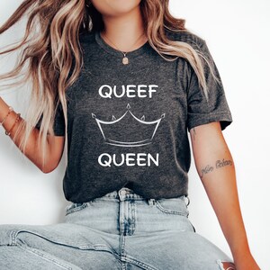 May include: Dark gray t-shirt with the words "QUEEF QUEEN" in white, along with a crown graphic. The shirt is a v-neck style and appears to be made of a soft material. The model is wearing blue jeans.