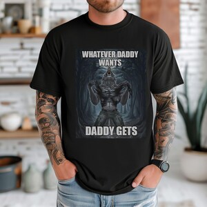 May include: Black t-shirt featuring a werewolf graphic in a shadowy forest setting. The text "WHATEVER DADDY WANTS" and "DADDY GETS" are printed above and below the image. The shirt is made of a soft material.