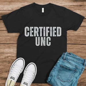 Pu&ograve; includere: Maglietta nera con il testo "CERTIFIED UNC" in bianco, indossata con jeans blu e scarpe da ginnastica bianche.