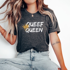Puede incluir: Una camiseta gris oscuro con el texto "Queef Queen" en blanco con una corona dorada sobre el texto. La persona que lleva la camiseta también lleva pantalones vaqueros azules.