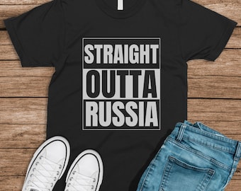 May include: Black t-shirt with white text that reads "STRAIGHT OUTTA RUSSIA". The shirt is on a wooden surface with a pair of white sneakers and blue jeans.