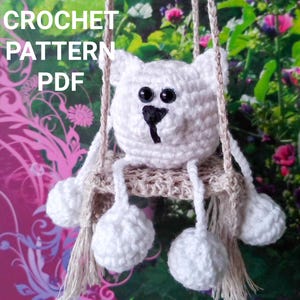 Pode incluir: Um urso branco de crochê com olhos pretos e um nariz preto está sentado em um balanço de crochê. O balanço é feito de fio bege e tem franjas na parte inferior. O urso está sentado em um assento de crochê bege.