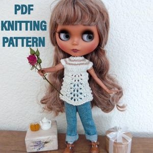 Può includere: Una bambola indossa un top bianco lavorato a maglia con un motivo traforato, jeans blu e stivali marroni. La bambola tiene in mano una rosa rosa. Il testo "PDF KNITTING PATTERN" è visibile in alto a sinistra.