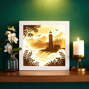 May include: A white framed, illuminated art piece depicting a lighthouse scene. The artwork features a lighthouse, mountains, and trees, with a warm, glowing light. A lit candle in a gold holder and a vase of white flowers are also present.