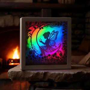 May include: A lighted fairy shadow box with a rainbow gradient background. The fairy is seated on a crescent moon, holding a star. The box is framed in white and sits on a stone surface. The background features flowers and foliage.