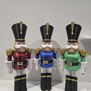 May include: Three toy nutcrackers in a row, each with a different coloured uniform. The nutcrackers have black hats with gold accents, white moustaches, and black boots. One is red, one is blue, and one is green.