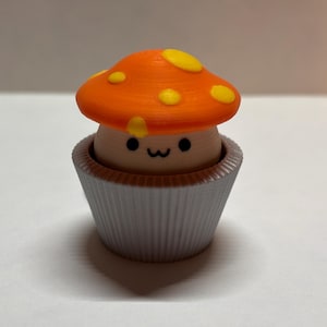 Op de afbeelding: Een 3D-geprint paddenstoelbeeldje met een oranje hoed en gele stippen, geplaatst in een grijze cupcake-vormige basis. De paddenstoel heeft een lachend gezicht. Het beeldje is een decoratief item.