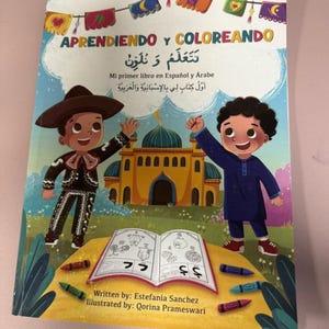 May include: A children's book titled "APRENDIENDO Y COLOREANDO" in Spanish and Arabic. The cover features illustrations of two children, one in a sombrero and the other in a blue outfit, with a mosque in the background. Crayons surround an open coloring book.