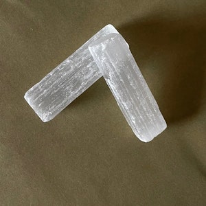 May include: Two white selenite crystal wands on a brown fabric background. The wands are smooth and have a striated texture.