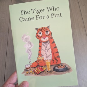 May include: A softcover book with a light green cover titled The Tiger Who Came For a Pint. The illustration features an orange tiger with black stripes holding a glass of beer, smoking a cigarette, with snacks and an ashtray on the table.
