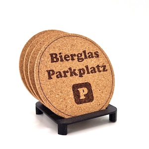 May include: Set of four cork coasters with the text "Bierglas Parkplatz" and a parking sign with the letter "P" in the center. The coasters are stacked and displayed on a black stand.