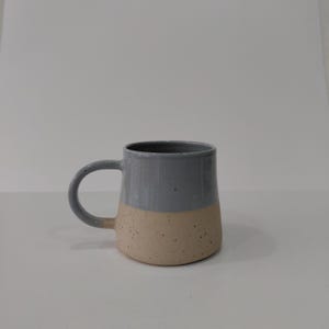 May include: A two-toned ceramic mug with a gray upper half and a speckled beige lower half. The mug has a rounded handle and a simple, modern design. The mug is suitable for coffee, tea, or other beverages.