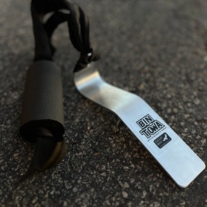 Puede incluir: Una herramienta de metal plateado con una pegatina negra que dice "Bin Toma con o sin un Tomahawk" y "Eagles Rest" con un logotipo de p&aacute;jaro. La herramienta est&aacute; sobre una superficie de asfalto gris. Tambi&eacute;n hay una correa negra con un mango de espuma negra en la imagen.