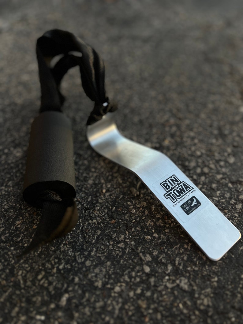 Puede incluir: Una herramienta de metal plateado con una pegatina negra que dice "Bin Toma con o sin un Tomahawk" y "Eagles Rest" con un logotipo de p&aacute;jaro. La herramienta est&aacute; sobre una superficie de asfalto gris. Tambi&eacute;n hay una correa negra con un mango de espuma negra en la imagen.