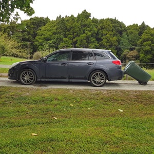 Puede incluir: Una camioneta gris oscuro con un contenedor de basura verde que se est&aacute; empujando hacia la parte trasera del coche. El coche est&aacute; aparcado en un camino de grava con hierba verde a ambos lados.