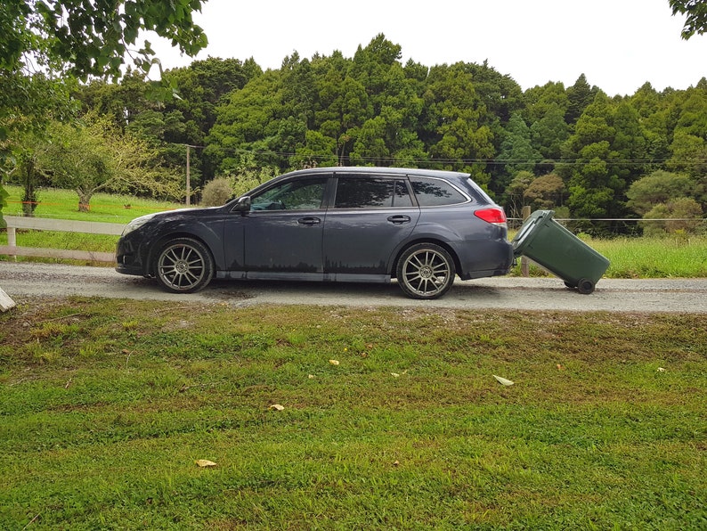 Puede incluir: Una camioneta gris oscuro con un contenedor de basura verde que se est&aacute; empujando hacia la parte trasera del coche. El coche est&aacute; aparcado en un camino de grava con hierba verde a ambos lados.