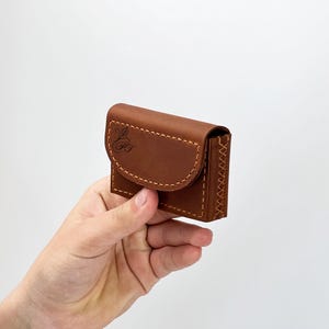 May include: A small, brown leather wallet with a curved flap and white stitching. The wallet has a decorative logo on the front and is held in a hand against a white background. The wallet appears to be new.