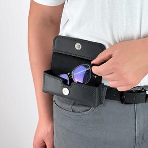 May include: Black leather sunglasses case attached to a belt. The case is open, revealing a pair of sunglasses with purple lenses. The case has a snap closure. The person is wearing a white t-shirt and gray pants.
