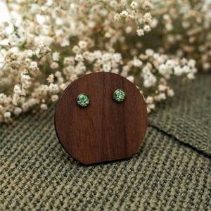 May include: A pair of round stud earrings with a green and white floral design, displayed on a dark wooden stand. The earrings are set against a backdrop of white flowers and a textured green fabric.