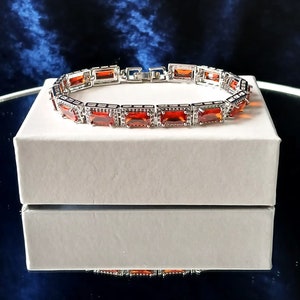 May include: A silver bracelet with a row of rectangular red gemstones. The bracelet is on a white box.