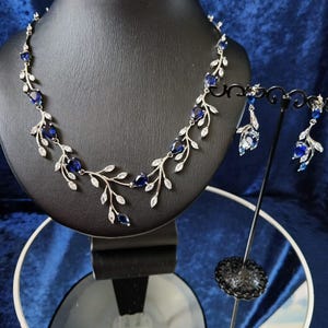 Puede incluir: Un collar y pendientes de plata con detalles de piedras preciosas azules. El collar presenta un delicado diseño de vid con hojas y piedras preciosas azules. Los pendientes son simples tachuelas con piedras preciosas azules.