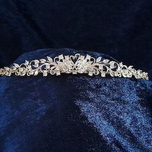 May include: A silver-toned tiara with a floral and leaf design, adorned with clear crystals. The tiara features a central motif and delicate, curving branches. The background is a dark blue, textured fabric.