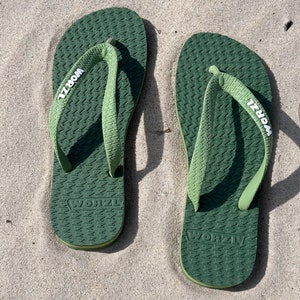May include: A pair of green flip-flops with the word "WORZTL" printed on the straps. The flip-flops are on a sandy surface.
