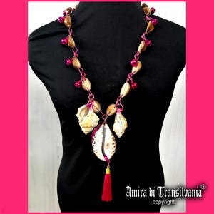 Può includere: Una collana artigianale con conchiglie naturali, perline marroni e dettagli rosa. La collana presenta un grande ciondolo centrale a conchiglia con un nappa rossa. Il design include il testo "Amira di Transilvania" e "copyright".