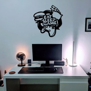 May include: A white desk with a black computer monitor, keyboard, and mouse. There is a black fan, a black speaker, and a small black figurine on the desk. A black metal wall decor with the text "Eat Sleep Game" is on the wall behind the desk. A small Christmas tree is in the corner of the room.