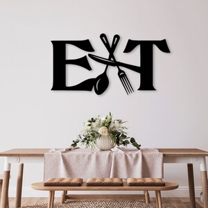 Puede incluir: Decoración de pared de metal negro con la palabra "EAT" en letras estilizadas. Un tenedor y una cuchara se cruzan detrás de la "T". La decoración está colgada en una pared blanca sobre una mesa de madera con un mantel blanco y un jarrón de flores blancas.