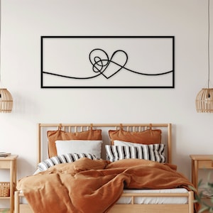 May include: A black metal wall art piece in the shape of a heart with a simple line design. The art is hanging above a bed with a brown comforter and pillows.