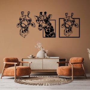 Op de afbeelding: Drie zwarte metalen wanddecoraties in de vorm van giraffenkoppen met verschillende uitdrukkingen. De giraffe links draagt een sjaal, de giraffe in het midden draagt een bril en de giraffe rechts draagt een sjaal. De decoratie is bevestigd aan een lichtbruine muur met twee bruine fauteuils en een houten tafel ervoor.