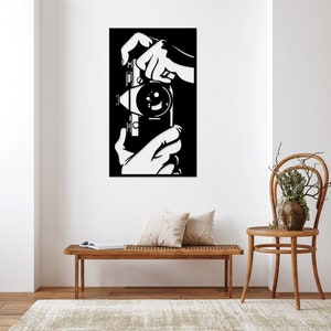 May include: Black and white wall art featuring a vintage camera. The artwork depicts hands holding the camera, with the lens and details of the camera body visible. The art piece is mounted on a white wall.