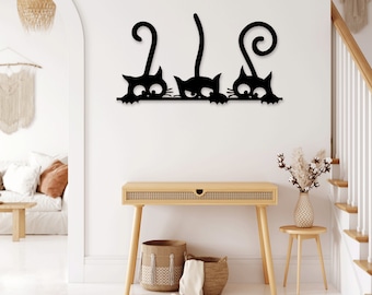 Op de afbeelding: Zwarte metalen wanddecoratie met drie gestileerde katten met hun staarten opgerold. De katten kijken naar de kijker en hebben een speelse uitdrukking.