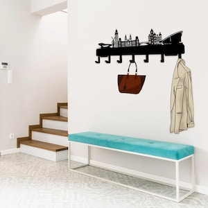 Puede incluir: Un perchero de metal negro con un diseño de ciudad, que incluye un bolso de mano marrón y una chaqueta beige colgados de ganchos. El perchero está montado en una pared blanca sobre un banco turquesa.